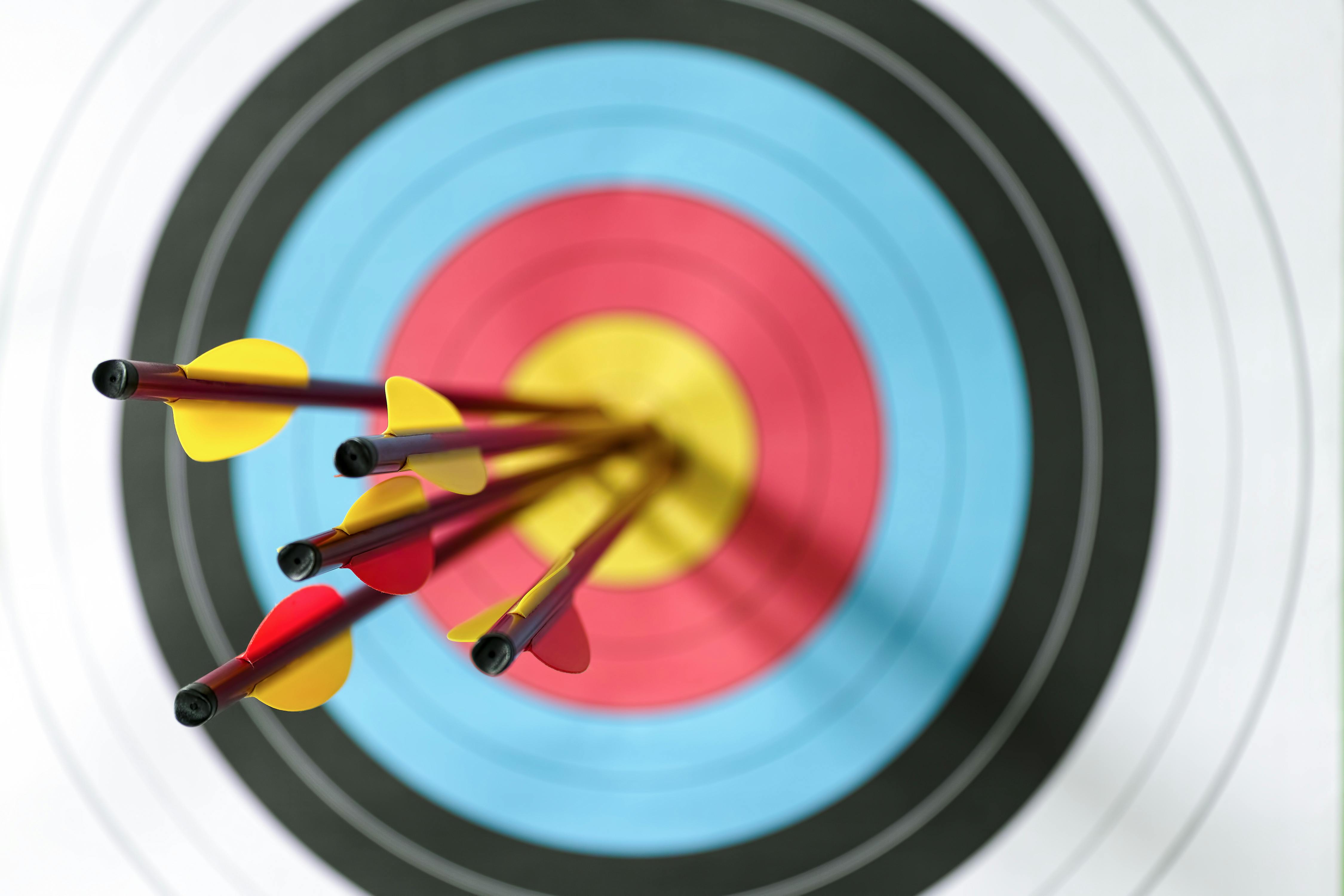 Five arrows in the bull's-eye of an archery target. Selective focus with the focus being on the back end of the arrow, with an out of focus target in the background.