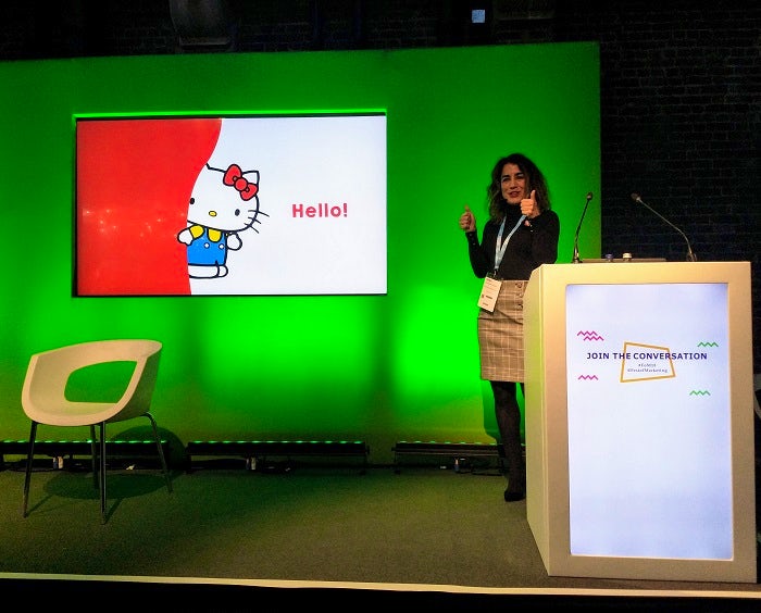 Martina Longueira gives a thumbs up on the Festival of Marketing content stage in front of a picture of Hello Kitty saying hello