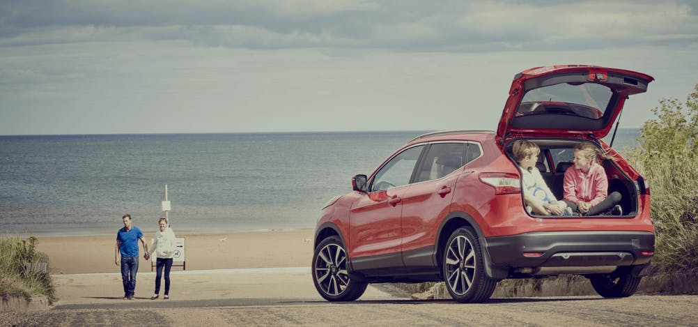 Red car on beach with children sitting in boot approached by mother and father