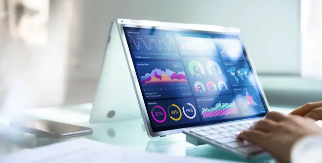 A pair of hands typing on the keyboard of a laptop displaying data visualisations on the screen.