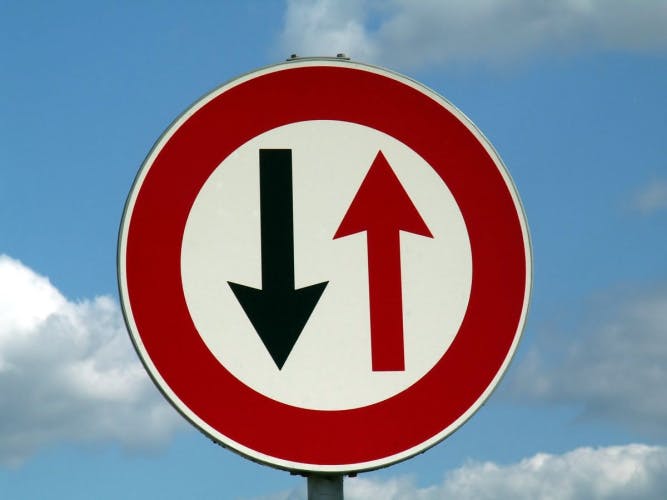 round road sign with red border with one black arrow pointing down and one red arrow pointing up on a blue sky background with clouds