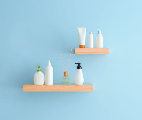 two wooden shelves, one short and one long, with the former above and to the right of the other, with white and clear bottles of assorted size on each, upon a light blue background