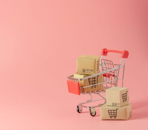 miniature shopping trolley containing two brown cardboard boxes, with similar boxes to its side, on a pink background