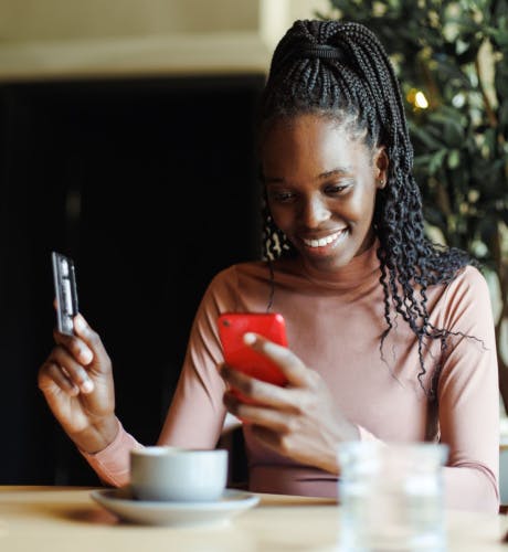 smiling black woman sitting at a table with a coffee cup holding a red smartphone in one hand and a credit card in the other. There is a black background over the woman's right shoulder and a plant over their left shoulder
