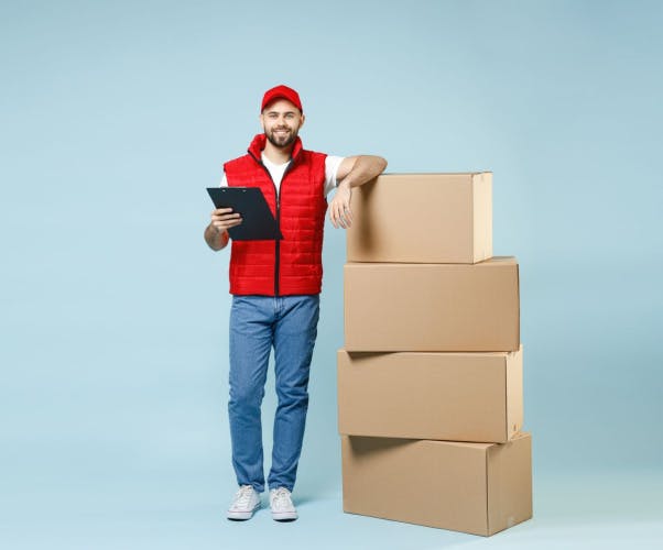 white man wearing red baseball cap, red gilet and blue jeans holding black clipboard resting elbow on a stack of brown cardboard boxes on a light blue background