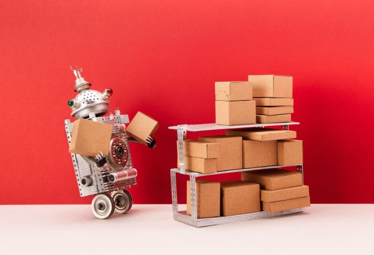 silver retro robot with wheels holding small brown cardboard boxes standing beside a unit of three shelves each carrying more cardboard boxes on a red background