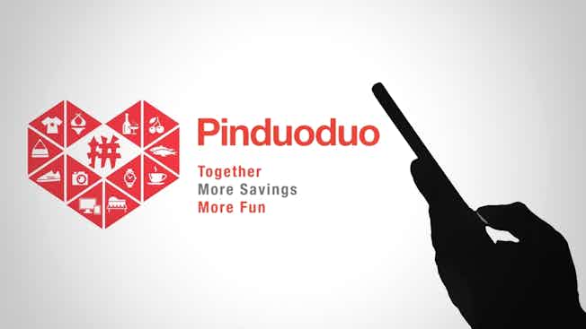 Pinduoduo's logo and its slogan, Together more savings more fun, are displayed on a wall with a hand holding a mobile phone silhouetted alongside it. 