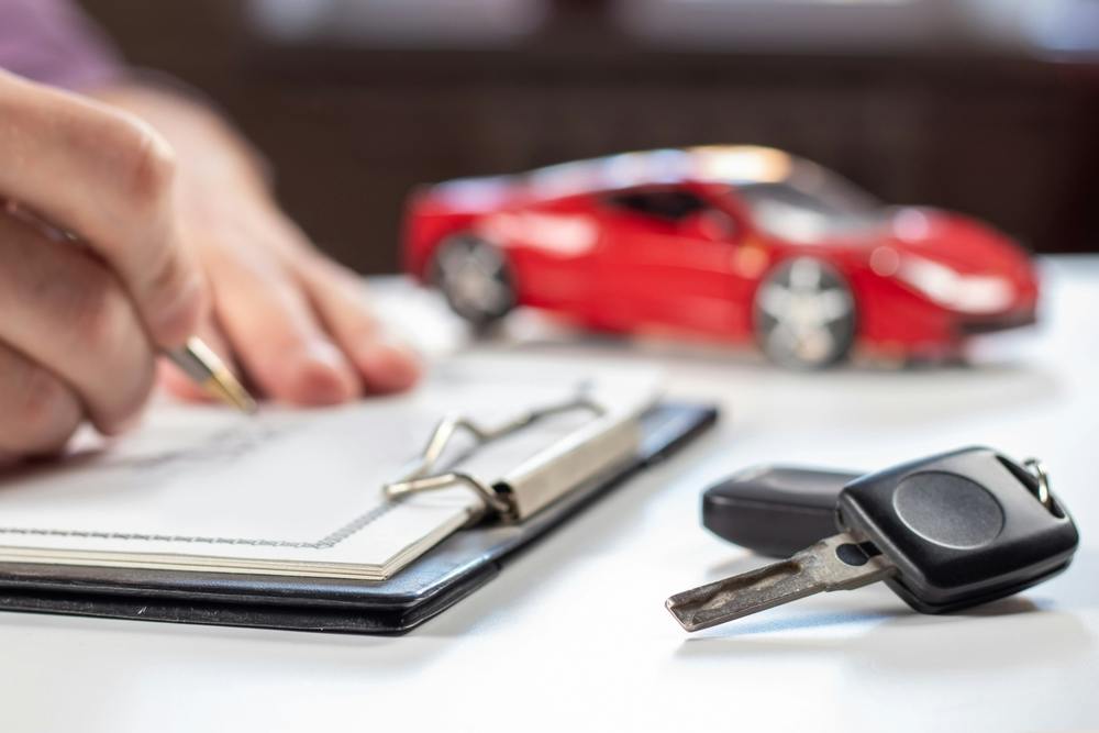 A person signs a document with a car key in the foreground and a red car in the background.